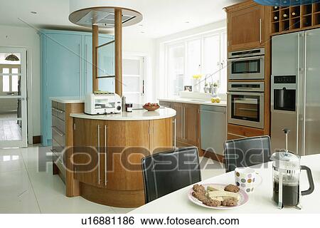 Stock Photograph - Cafetiere and biscuits on white table in large modern kitchen with American-style fridge freezer and island unit. Fotosearch