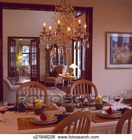 Stock Photograph - Candles on table set for dinner below glass chandelier in traditional diningroom with double doorway open to livingroom. Fotosearch