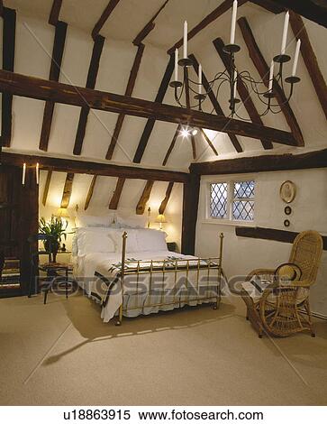 Cane armchair and brass bed in beamed country bedroom with cream carpet and candelabra and apex ceiling View Large Photo Image Stock Photography - Cane armchair and brass bed in beamed country bedroom with cream carpet and candelabra and apex ceiling . Fotosearch