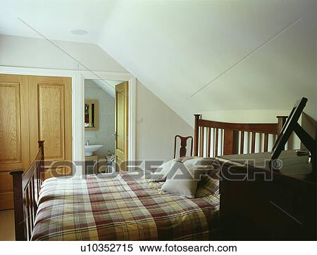 Stock Photography - Checked bedlinen on wooden bed in white attic bedroom with door open to ensuite bathroom. Fotosearch