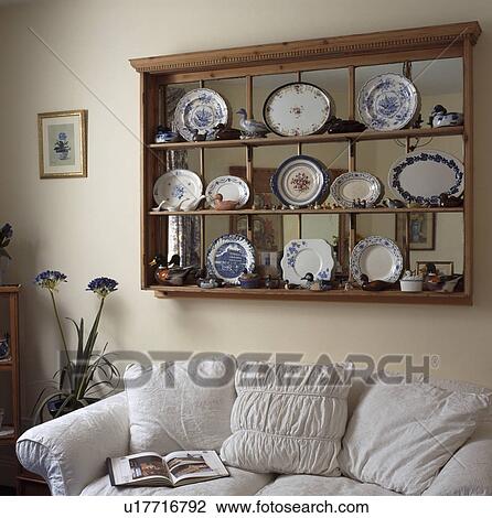 Stock Image - China plates on pine shelves over internal window above white sofa in living room. Fotosearch