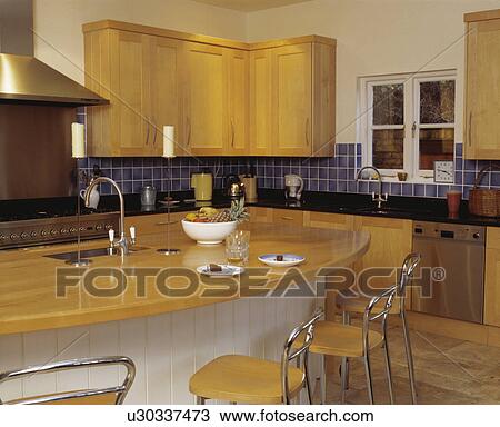 Stock Image - Chrome and wood stools at breakfast bar with wood worktop in modern kitchen with fitted wooden cupboards. Fotosearch