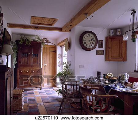 Stock Image - Circular clock on wall in white cottage dining room with tiled floor and large cupboard. Fotosearch