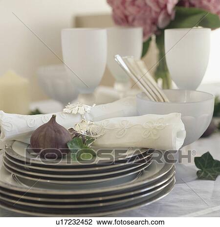 Stock Image - Close-up of fresh fig and embroidered napkin on stack of white plates with grey edging. Fotosearch