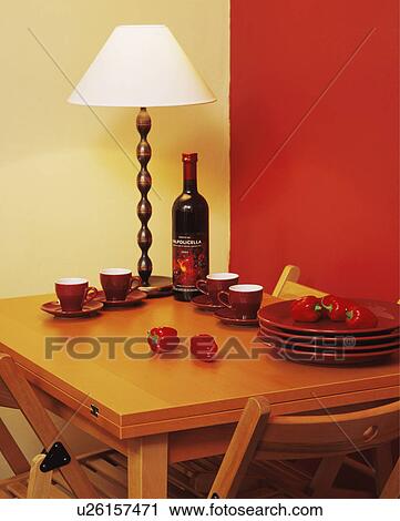 Stock Image - Close-up of lighted lamp with red crockery on wooden table in red and neutral dining room. Fotosearch