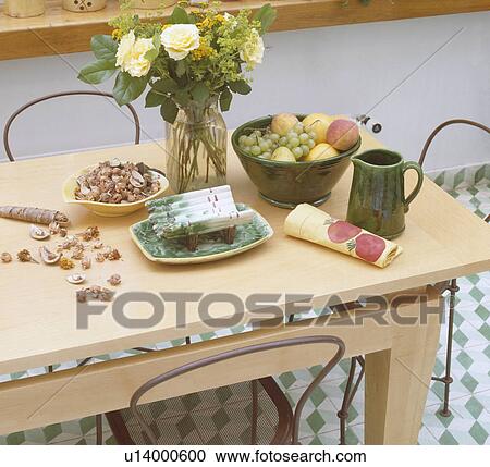 Stock Image - Close up of roses in vase and fruit in green bowl with green pottery jug on pale wood table. Fotosearch