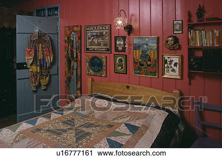 Stock Image - Colourful pictures on panelled pink wall above pine bed with patchwork quilt in sixties cottage bedroom. Fotosearch