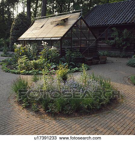 Stock Image - Country garden with circular bed in brick paving with greenhouse in background. Fotosearch