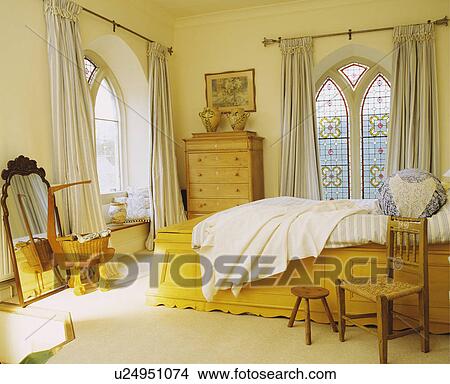 Picture - Cream curtains at stained glass Gothic windows in cream bedroom with bed on raised wooden yellow base. Fotosearch