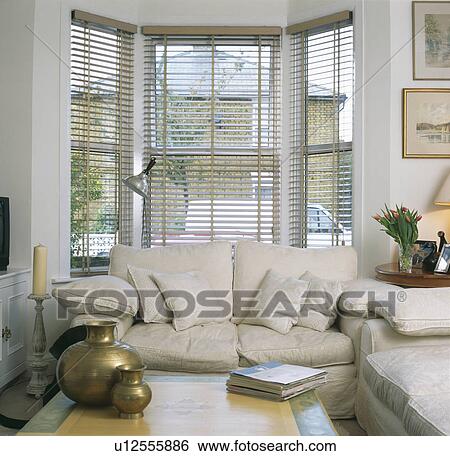 Cream Sofa In Front Of Bay Window With Slatted Blind In Living Room With Brass Pots On Coffee Table Stock Photograph