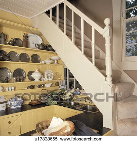 Stock Photography - Double steel sink below staircase in small yellow understairs kitchen with pewter plates and white china on shelves. Fotosearch