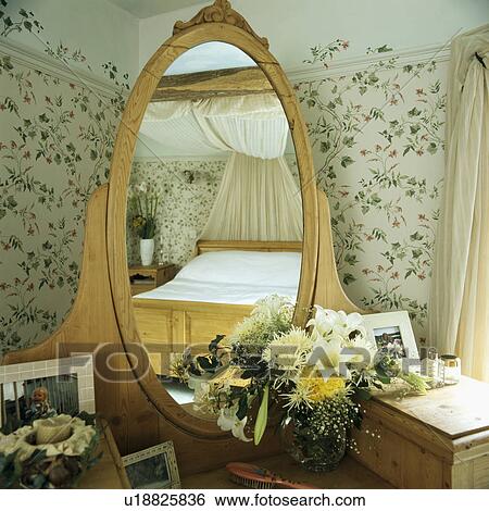 Dressing table with fresh flowers and integral oval mirror reflecting pine bed in country bedroom with floral wallpaper View Large Photo Image Stock Photograph - Dressing table with fresh flowers and integral oval mirror reflecting pine bed in country bedroom with floral wallpaper. Fotosearch
