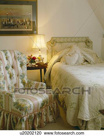 Stock Image - Floral and pink striped armchair in attic bedroom with lighted lamp beside single bed with upholstered headboard and matching quilt. Fotosearch