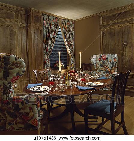 Stock Photograph - Floral armchairs and antique table set for dinner in panelled dining room with floral curtains. Fotosearch