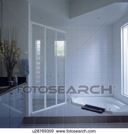 Glass shower cabinet and sunken corner bath in modern white tiled bathroom View Large Photo Image Stock Photo - Glass shower cabinet and sunken corner bath in modern white tiled bathroom. Fotosearch