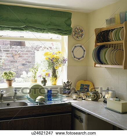Stock Photo - Green blind above stainless steel sink with drainer in pastel yellow kitchen with pine plate rack on the wall. Fotosearch