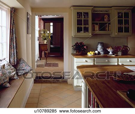 Stock Photography - Island unit with wooden worktop in traditional cottage kitchen with terracotta tiled floor and cushions on windowseat. Fotosearch