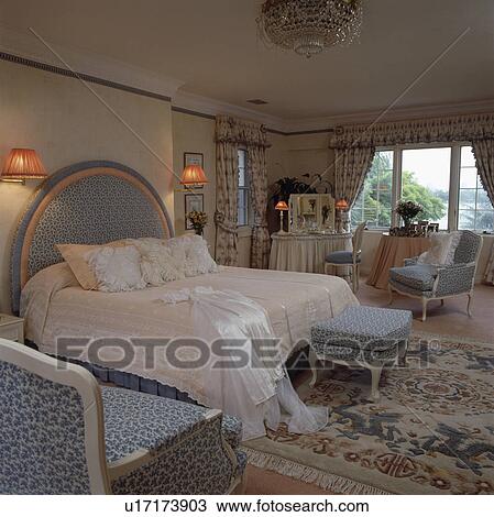 Stock Image - Lace pillows on bed with upholstered headboard to match chairs and stool in traditional bedroom with floral rug. Fotosearch