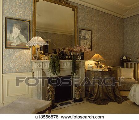 Large Mirrror Above Fireplace In Bedroom With Patterned Wallpaper And Lamp On Brown Cloth On Circular Table Stock Photograph