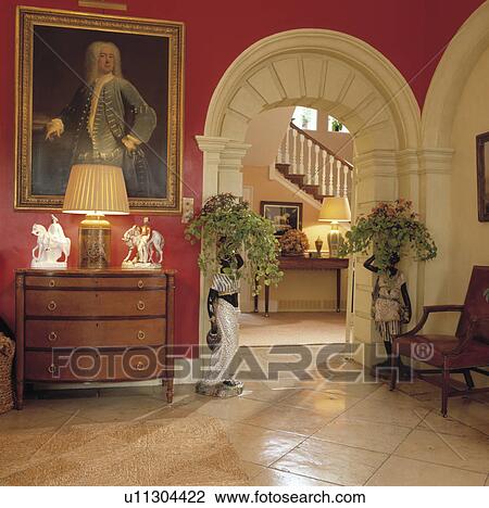 Stock Image - Large oil painting above chest of drawers in red Georgian hall with stone floor and white arch with view of staircase. Fotosearch