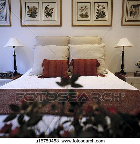 Stock Image - Large white bedroom with framed pictures on wall and red and white cushions on bed. Fotosearch