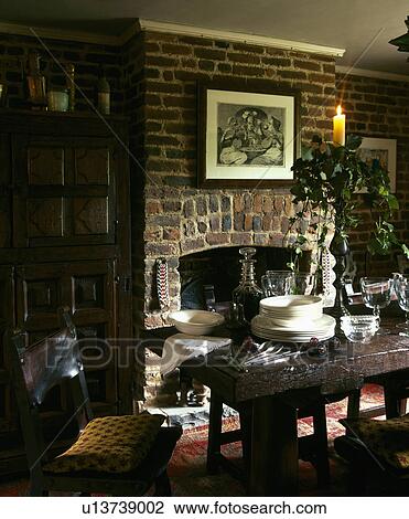 Stock Image - Lighted candle in tall candlestick on oak table in country dining room with fireplace in exposed brick wall. Fotosearch