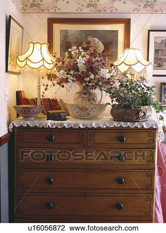 Stock Image - Lighted lamps with fresh flowers and houseplant on antique chest of drawers with lace cloth. Fotosearch