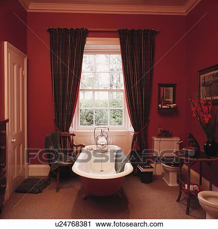 Stock Image - Long curtains on either side of sash window above freestanding clawfoot bath in red bathroom with beige carpet. Fotosearch