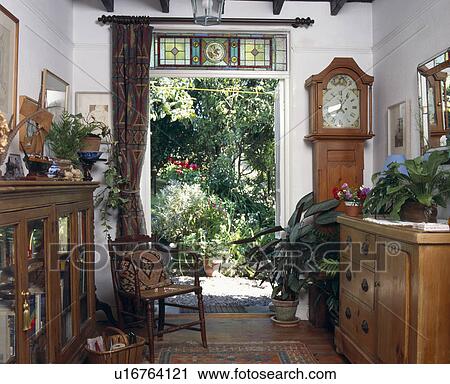 Stock Image - Longcase clock in hall with stained glass window above open door with view of the garden. Fotosearch