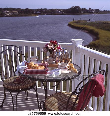 Stock Photo - Modern metal chairs and circular glass-topped table laid for breakfast with croissants and coffee on balcony overlooking wide river. Fotosearch