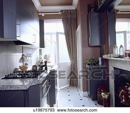 Stock Image - Narrow galley kitchen with grey fitted cupboards and white+black tiled floor and beige curtain at open door. Fotosearch