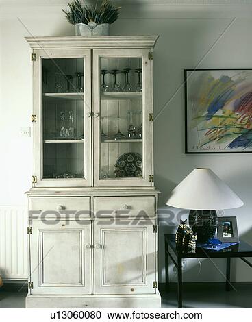 Stock Image - Narrow grey dresser with glazed doors beside table with white lamp. Fotosearch