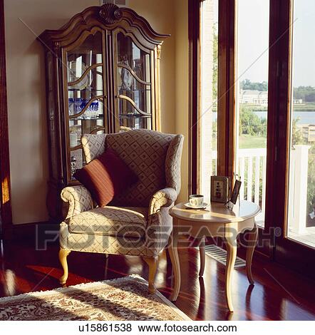 Neutral patterned armchair in front of antique glass-fronted bookcase in corner of livingroom with patio doors View Large Photo Image Stock Photo - Neutral patterned armchair in front of antique glass-fronted bookcase in corner of livingroom with patio doors. Fotosearch