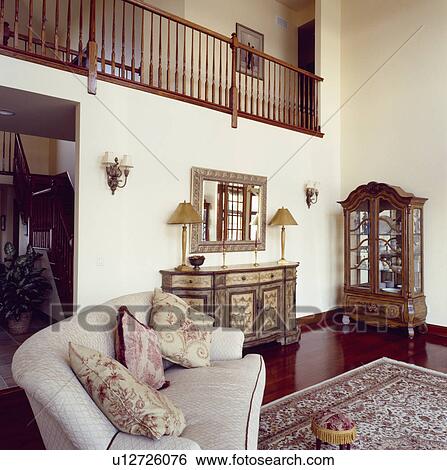 Stock Photograph - Neutral sofa with toile-de-Jouy cushions and painted antique sideboard and glass-fronted cupboard in modern double-storey livingroom. Fotosearch