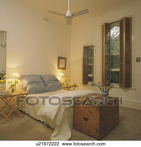 Stock Image - Old pine chest below bed with blue pillows and cream quilt and linen in white bedroom with wooden plantation shutters. Fotosearch