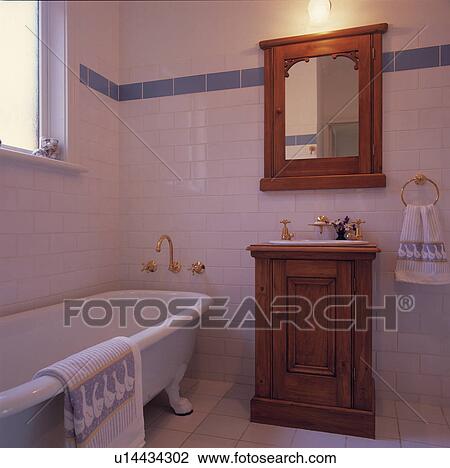 Stock Image - Old pine mirror above pine cabinet with basin in white tiled bathroom with rolltop bath. Fotosearch