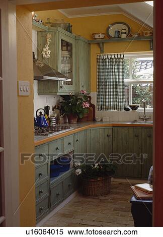 Stock Photography - Pastel green cupboards and units in small pale orange kitchen extension with green checked curtains. Fotosearch