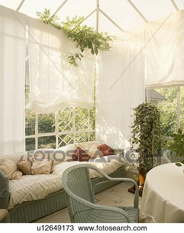 Stock Image - Pastel green wicker chair and sofa with white cushions in conservatory dining room with houseplants and white blinds. Fotosearch