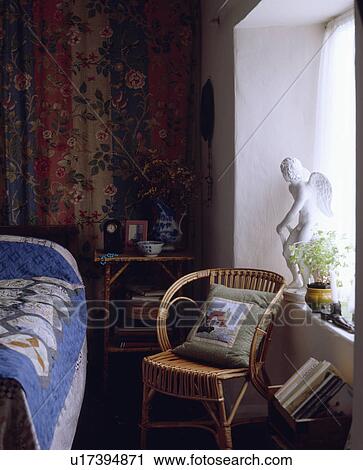 Patchwork cushion on wicker chair beside statue on windowsill in small cottage bedroom with drapes behind bed View Large Photo Image Stock Image - Patchwork cushion on wicker chair beside statue on windowsill in small cottage bedroom with drapes behind bed. Fotosearch