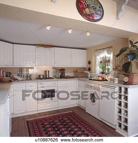 Stock Photograph - Patterned rug in small white kitchen extension with washing machine and dishwasher. Fotosearch