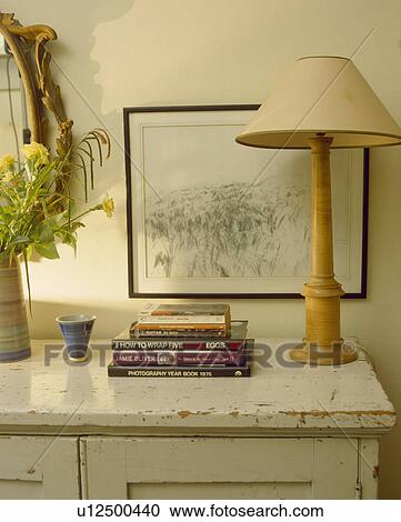 Stock Image - Picture above pile of books and lamp with wooden base and cream shade on cream painted cupboard . Fotosearch