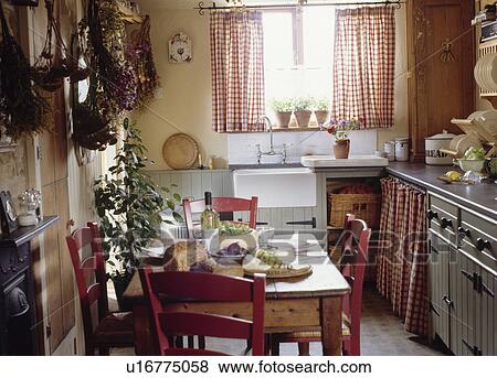 Stock Photo - Pine table and red chairs in cottage kitchen with red checked curtains at the window over the sink. Fotosearch