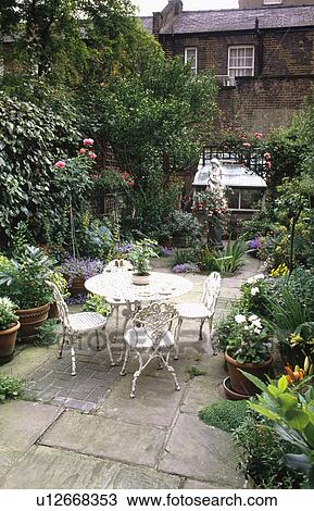Stock Image - Plants in pots & white metal table and chairs on stone paved patio in town garden with statue and greenhouse. Fotosearch