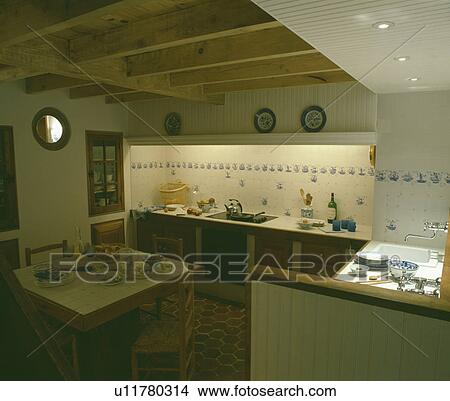 Picture - Recessed lighting and wall tiles above fitted cupboards in small white kitchen with wooden table with white tiled top. Fotosearch