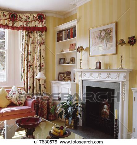 Stock Photography - Red trimmed pelmet on floral curtains in traditional sittingroom with yellow striped wallpaper and white fireplace. Fotosearch