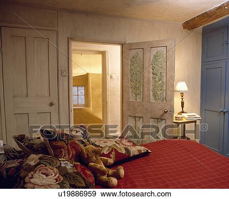 Rose-patterned tapestry cushions on red bedcover in country bedroom with painted trees on open door beside lighted lamp View Large Photo Image Stock Photo - Rose-patterned tapestry cushions on red bedcover in country bedroom with painted trees on open door beside lighted lamp. Fotosearch