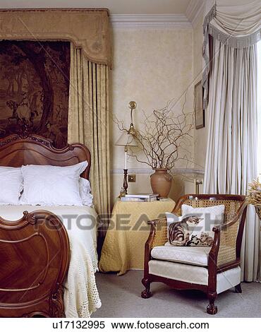 Silk Drapes Above Mahogany Bed In Neutral Bedroom With Bergere