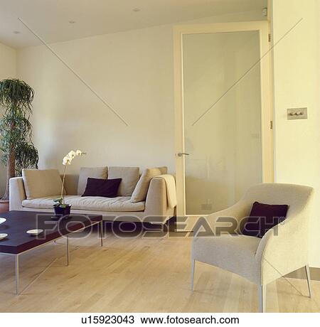 Stock Image - Small cream armchair and beige sofa in modern livingroom with glass door and maple wood flooring. Fotosearch