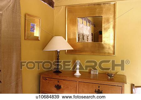 Stock Photo - Square gilt mirror above chest-of-drawers with white lamp. Fotosearch