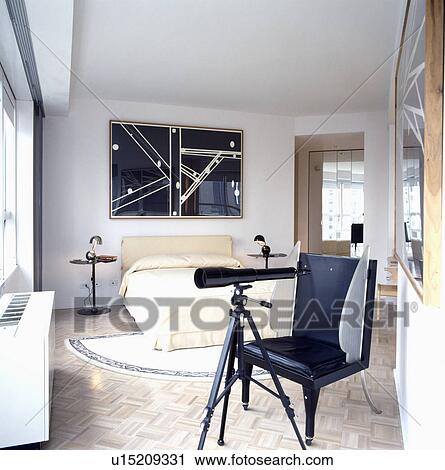 Stock Image - Telescope with black leather chair in modern white bedroom with black+white abstract picture above bed with cream linen. Fotosearch